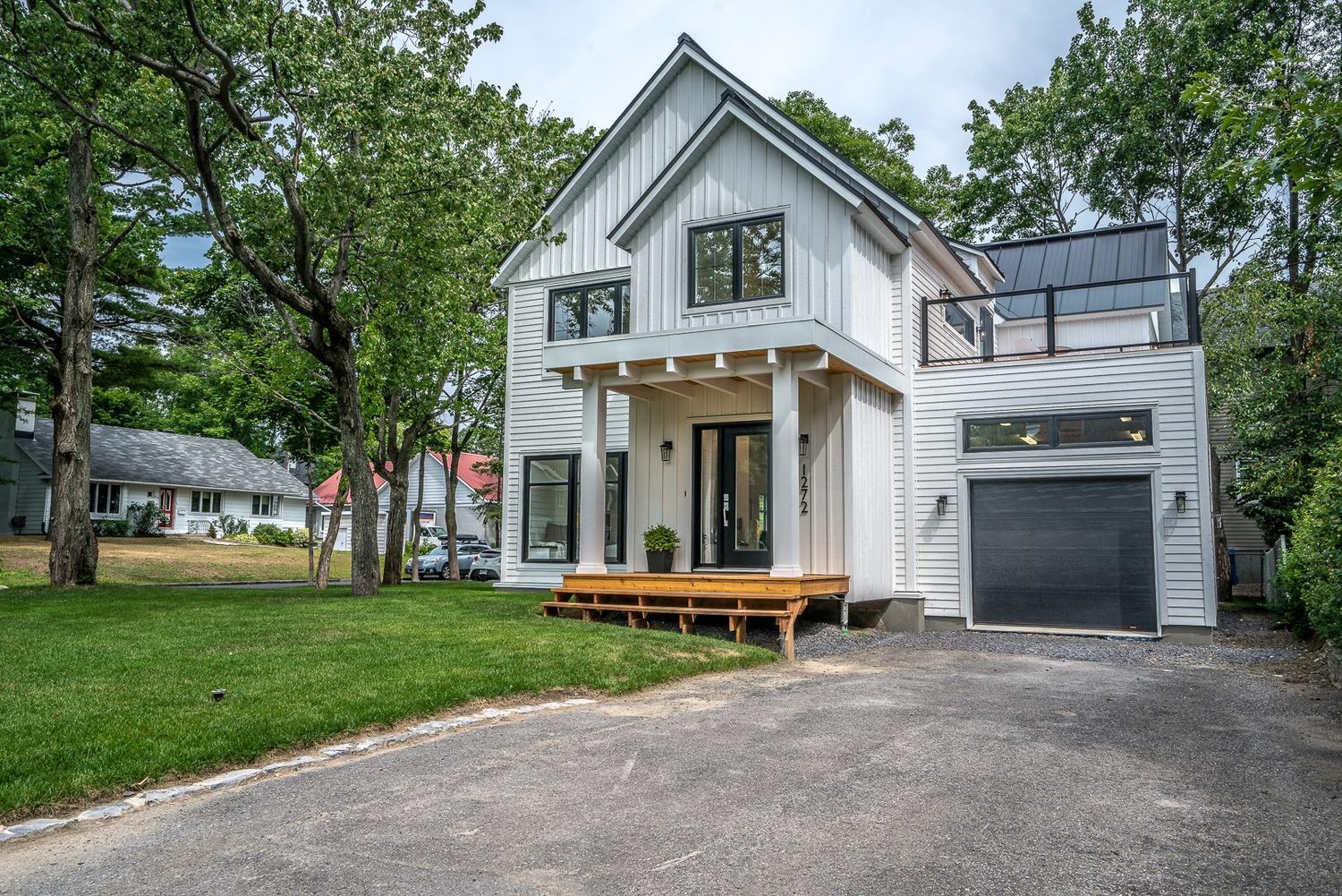 Une maison blanche à toit de tôle, lucarne, fenêtres noires, garage et terrasse de bois