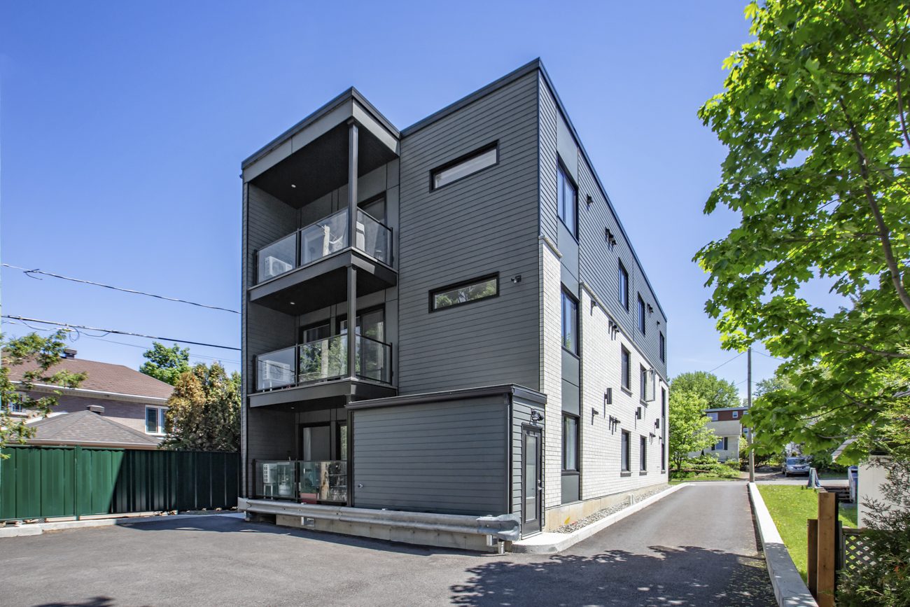 Un immeuble locatif gris à trois étages, balcons vitrés, fenêtres noires, sous les arbres