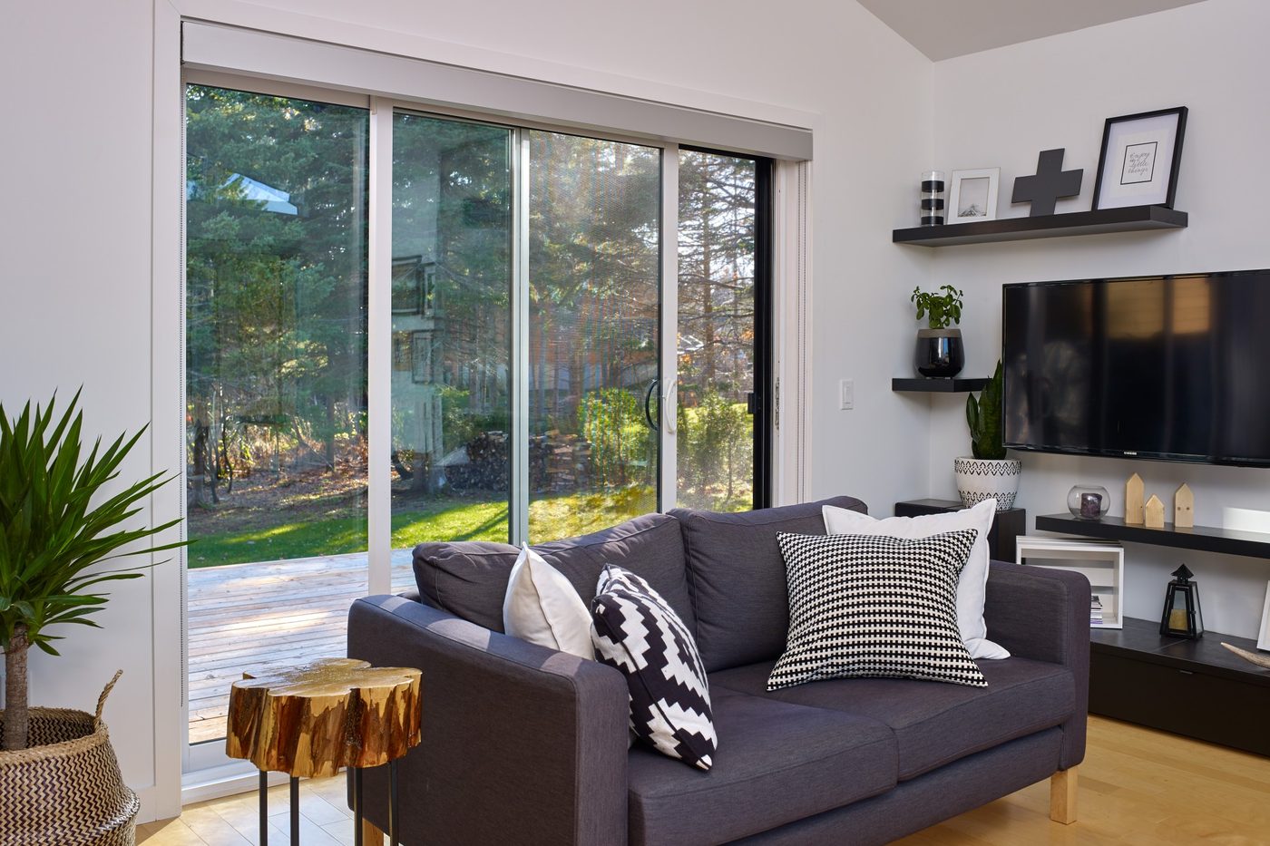 Une porte-patio blanche ouverte sur une terrasse boisée, un divan gris au premier plan