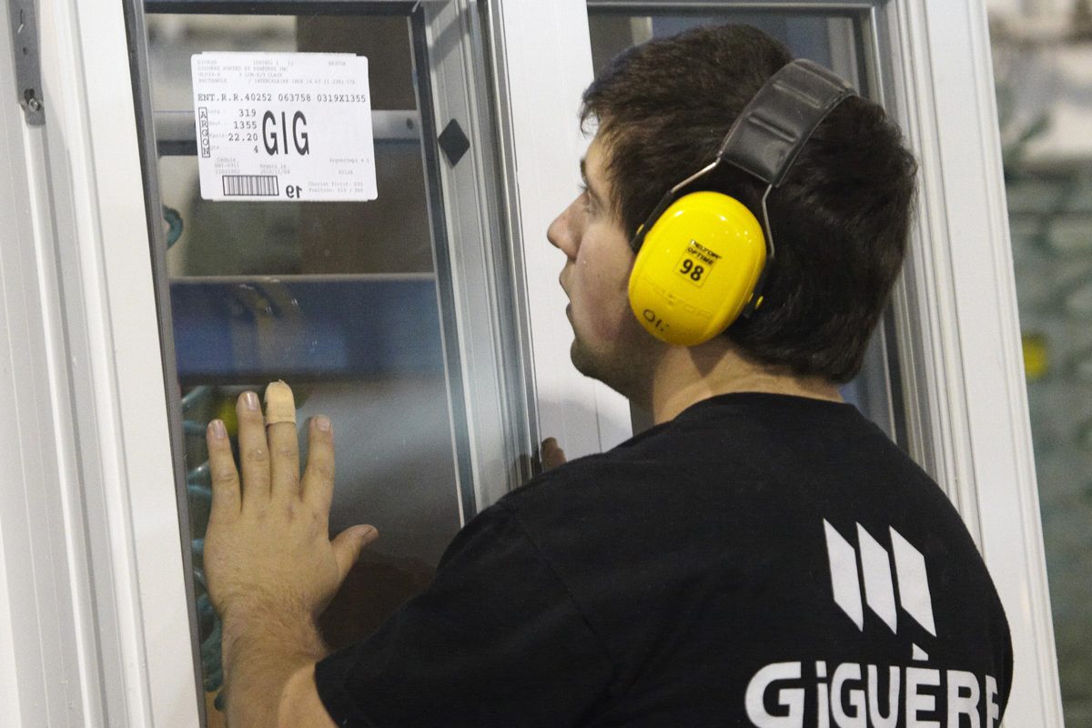 Un employé de Giguère, chandail noir au logo, protège-oreilles jaunes, manipule une fenêtre à l'usine